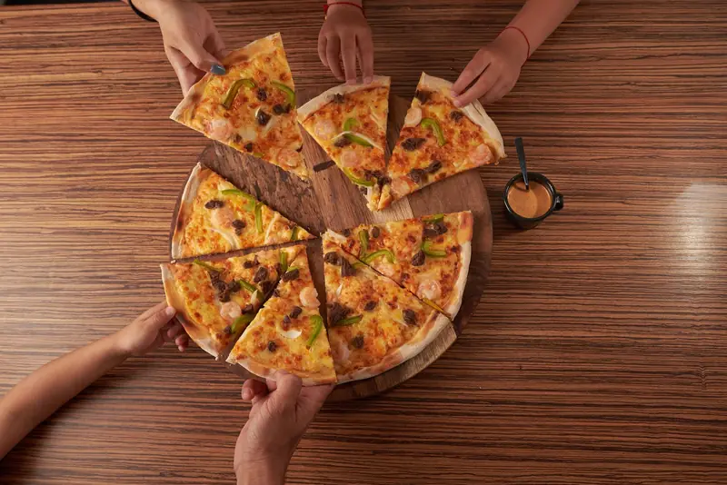 Top view of people sharing a cheesy pizza with various toppings on a wooden table