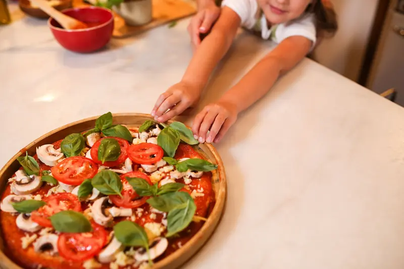 A child reaches for a homemade pizza with fresh toppings on a kitchen counter