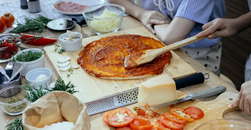 From above side view of crop anonymous women smearing tomato sauce by spatula over dough base while preparing pizza together in kitchen
