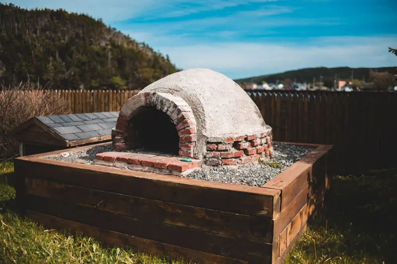 Aged outdoor pizza oven on wooden construction on grass behind mountains under blue cloudy sky